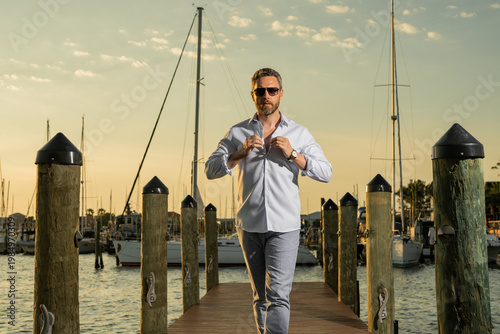Rich fashion business man dreaming and thinking of yacht. Well-dressed fashion man in fashionable eyeglasses and shirt posing near yacht. Male model in yacht club. Portrait of stylish man. Male