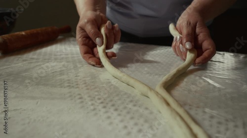 Female baker skillfully twists and shapes dough into a braided form on a textured surface, with a rolling pin and bowl visible in the background