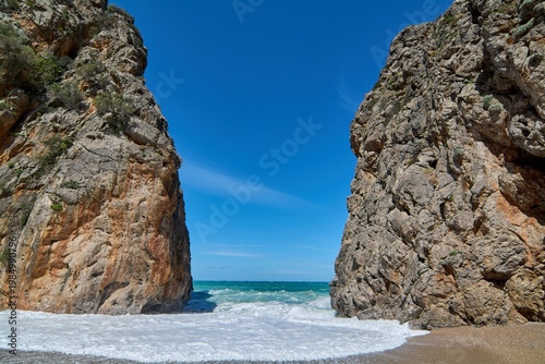 Dramatic coastal cliffs framing the ocean horizon with splashing waves on a pebble beach under a bright sunny day for travel marketing