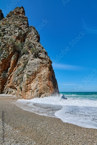 Wild pebble beach with vertical cliff and turquoise sea water. Natural coastal landscape with splashing waves and sunny sky. Perfect for vacation and nature advertising campaigns. Copy space.