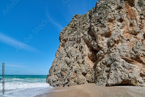 Scenic horizontal view of a steep cliff on a natural beach with sea foam. Wild coastal environment for tourism marketing and Mediterranean vacation concepts. High quality commercial landscape.