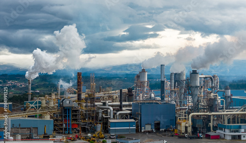 Aerial view of industrial complex billowing smoke into the cloudy sky, a stark contrast against the distant hills, Medellin, Colombia.