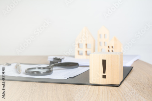 Single exclamation mark on a wooden block. Blurred background with miniature houses, magnifying glass and contract. Real estate warning concept.