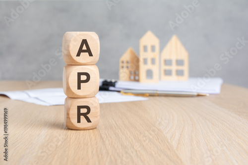 APR - Annual Percentage Rate - acronym on wooden blocks. Blurred background with miniature houses and documents. Mortgage loan concept.
