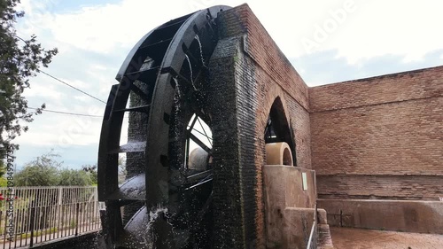Ancient large black metal water wheel beside historic brick building in Spanish countryside sunny day traditional irrigation heritage landmark rural architecture in murcia