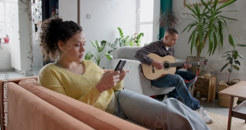 Young Adult Playing Guitar, Learning Music, Roommate Looking at Phone on Sofa
