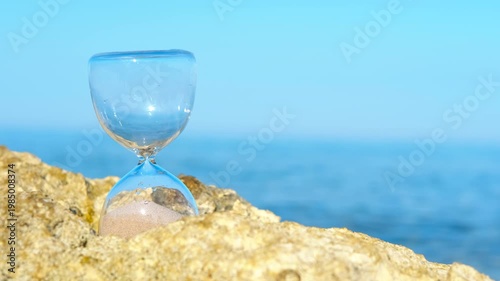 Hourglass measuring time on a rock by the sea. Crystal clear hourglass with sand pouring through it sits on a coastal rock, measuring the passage of time against a serene blue ocean