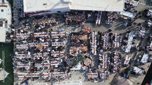 Aerial drone view of Easter celebration in Naousa village on Paros island, Greece, showing people gathered at outdoor tables in a festive Mediterranean atmosphere. Ideal for culture, travel, holidays