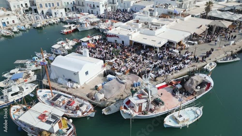 Aerial drone view of Easter celebration in Naousa village on Paros island, Greece, showing people gathered at outdoor tables in a festive Mediterranean atmosphere. Ideal for culture, travel, holidays