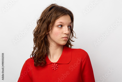 Bilde på lerret Young woman with curly brown hair wearing a red sweater looks to the side with a neutral expression on a plain white background