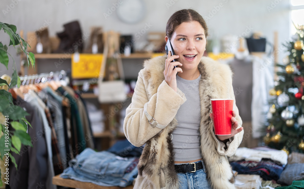 Fototapeta premium European girl with cup of coffee talking on phone in womens clothing store. Shopping as hobby and pastime. Megamall, sales and promotions, offline store of branded clothing manufacturer