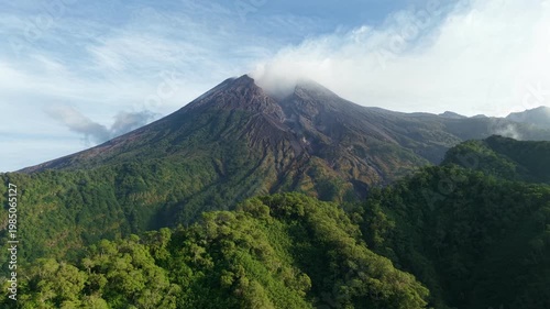 An aerial view of Mount Merapi surrounded by dense forest in Java, Indonesia. A view of volcanic terrain and hills under a clear blue sky.