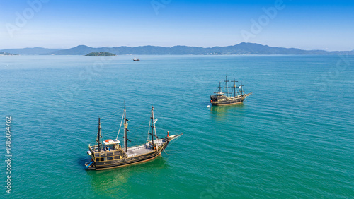 Fotografi Two tall-ship pirate vessels anchored on the turquoise waters of Florianopolis bay under clear blue sky
