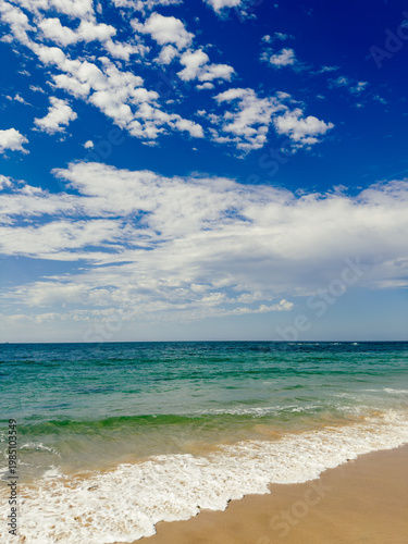 Ocean waves washing up to a sandy beach under a blue sky with light white clouds
