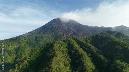 Mount Merapi in Indonesia, a majestic active volcano towering above lush tropical rainforest with rolling clouds.