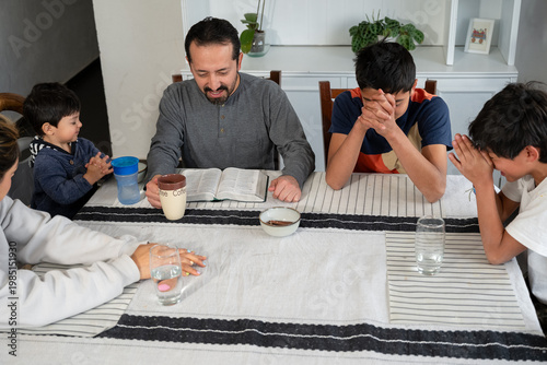 Hispanic family bowing heads in prayer after reading scripture at home