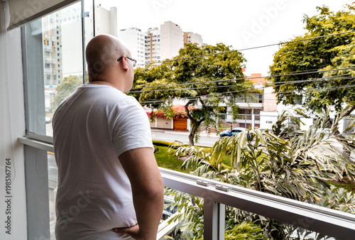 Bald hispanic man standing by window looking at city view