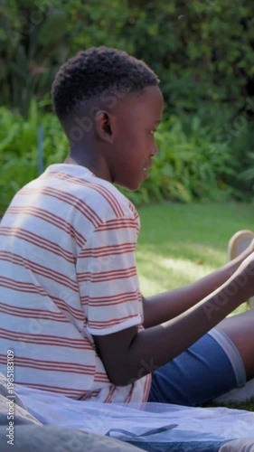 Vertical video: Checking tin cup, boy in striped T glancing off-frame drinking on blanket in garden