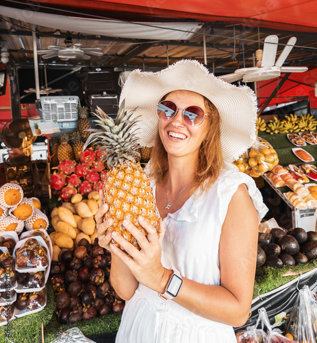 Tourist woman selecting fresh pineapple at colorful street market. Tropical lifestyle, organic food choice and cheerful summer atmosphere with variety of exotic fruits