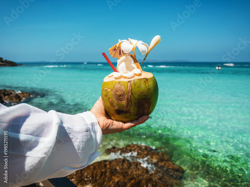 Hand holding exotic coconut cocktail hand holds exotic coconut cocktail with lilawadee flower straw, on sea tropical beach Crystal clear ocean blue sky create refreshing summer vibe travel inspiration