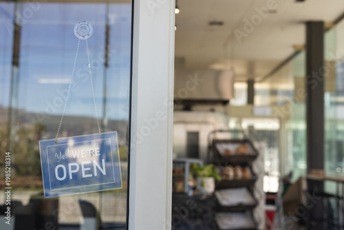 Yes WE'RE OPEN sign is hanging on glass door with dotted chain, blurred bakery interior