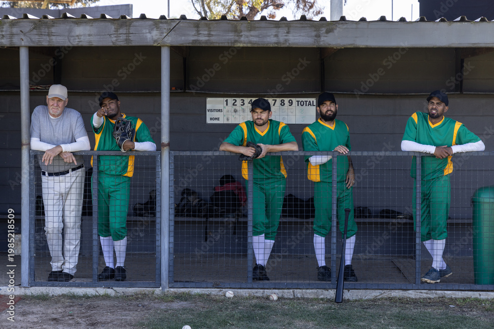 Fototapeta premium Diverse male baseball team and coach standing on dugout rail in green uniforms holding gloves, bat