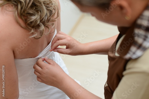 bridesmaid hands fastening the back of a white wedding dress, close up of bride getting dressed for her wedding ceremony, woman helping bride to zip up her bridal gown, wedding morning preparation det
