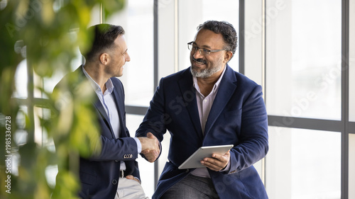 Obraz na płótnie Two smiling businessmen sit together in office lounge, expressing mutual respect and shaking hands during meeting while holding digital tablet
