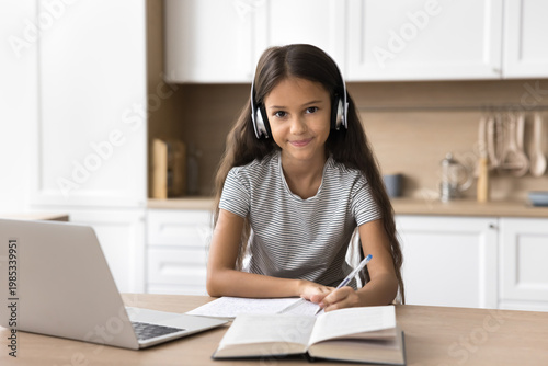 Cuadro en lienzo Portrait of cute little 7s schoolgirl studying at home using laptop, wear headphones listening audio course, improve english knowledge via application or educational platform