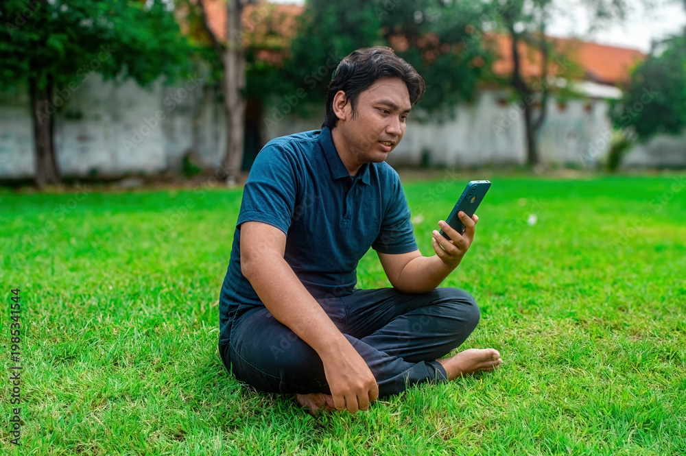 Obraz premium A man is sitting cross legged on a grassy lawn, holding a smartphone and seemingly engaged in a video call. The background features trees and a wall, suggesting an outdoor setting