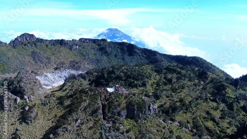 Cinematic dolly out drone shot reveals the rugged Puncak Sejati of Mount Sumbing, showcasing hikers gathered at the summit under a clear blue sky