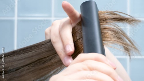 Woman straightening her hair with a hair straightener against background of blue tiles in bathroom.