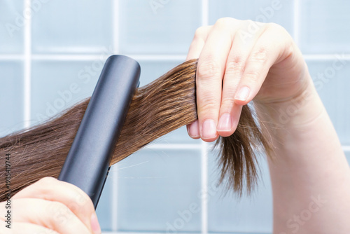 Woman straightening her hair with a hair straightener against background of blue tiles in bathroom.