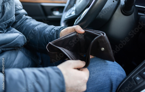 Man sitting in car at gas station during fuel crisis,checking empty wallet and holding payment card.Concept of rising fuel prices.Driver leaning on steering wheel.Close of fuel nozzle into car tank.