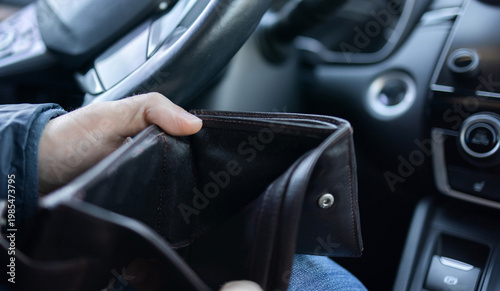 Man sitting in car at gas station during fuel crisis,checking empty wallet and holding payment card.Concept of rising fuel prices.Driver leaning on steering wheel.Close of fuel nozzle into car tank.