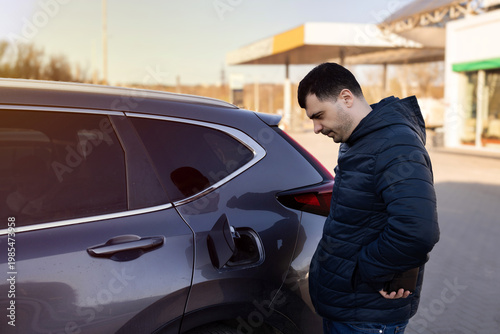 Man sitting in car at gas station during fuel crisis,checking empty wallet and holding payment card.Concept of rising fuel prices.Driver leaning on steering wheel.Close of fuel nozzle into car tank.