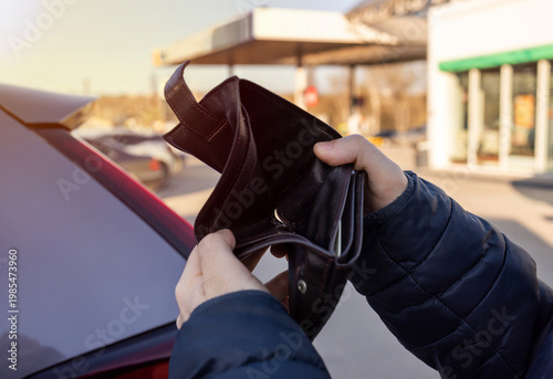 Man sitting in car at gas station during fuel crisis,checking empty wallet and holding payment card.Concept of rising fuel prices.Driver leaning on steering wheel.Close of fuel nozzle into car tank.