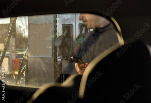 Man sitting in car at gas station during fuel crisis,checking empty wallet and holding payment card.Concept of rising fuel prices.Driver leaning on steering wheel.Close of fuel nozzle into car tank.
