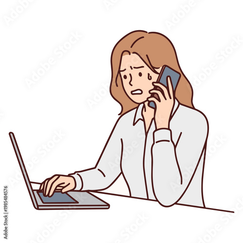 A woman sits at a desk looking stressed while talking on her phone and typing on a laptop.