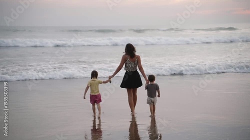 Back view of mother walking with her son and daughter along the beach at sunset, enjoy family time. Golden hour light, scenic ocean views. Concept of bonding, summer vacation, and outdoor lifestyle