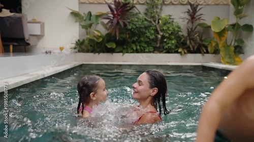 Joyful family pool scene mother hugging and playfully tossing her daughter over the water while boy jumps into the pool with big splashes. They laughter, carefree summer moments