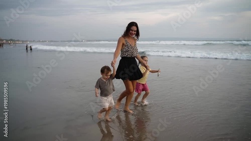 Mother running with her son and daughter toward the ocean along a sandy beach at sunset. Concept of family together moment in golden hour light on vacation, carefree play. 