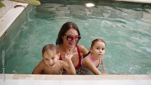 Mother chills with her children in the pool, all looking at the camera, smiling and laughing in a joyful family moment. Family vacation at tropical summer villa
