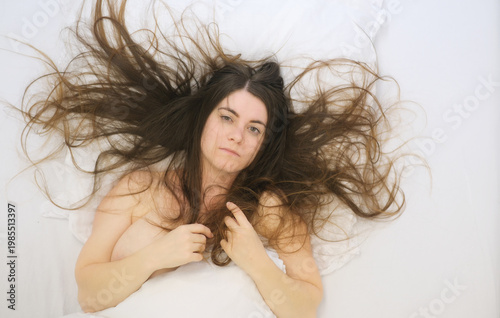 Young woman lying on white bed with long flowing hair spread around head looking calmly at camera expressing intimacy vulnerability softness rest and quiet emotional openness