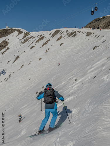 Skier dropping off piste to follow another skier in French Alps le plagne. Skier moving a speed down hill in blue ski suit and black backpack, set against blue bird skies.