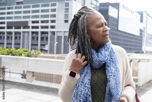 Senior African American woman touching hair on terrace by railing, wearing smartwatch, shoulder bag