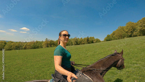 Fotografie PORTRAIT: Cheerful young horsewoman horseback riding through lush green nature