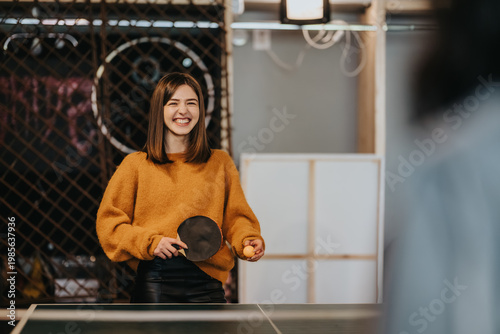 A smiling young woman plays table tennis holding a paddle and ball. She enjoys a casual indoor game while a friend watches nearby.