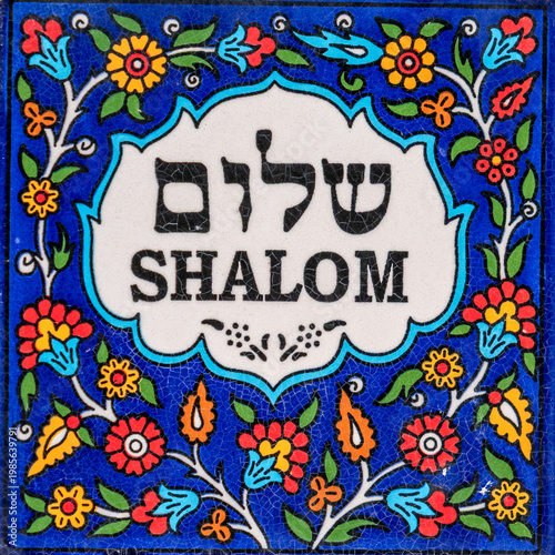 Colorful Armenian tile that says Shalom in English and Hebrew text on white with a flowered blue background.
