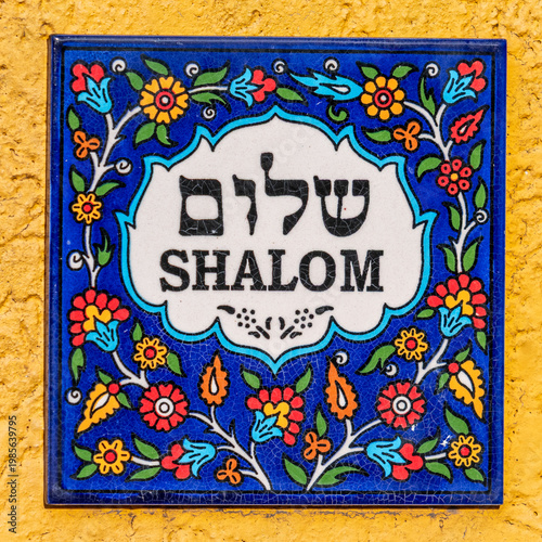 Colorful Armenian tile that says Shalom in English and Hebrew text on white with a flowered blue background against a yellow wall.
